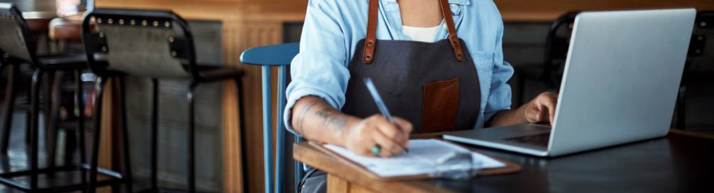 Restaurant manager using a laptop and making notes on a clipboard at a bar